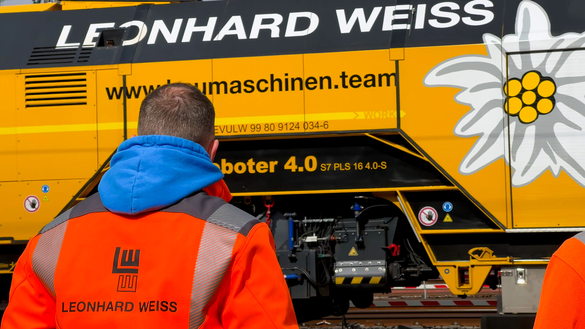 Ein Arbeiter in leuchtend orangefarbener Kleidung steht vor einer gelben Maschine mit dem Logo von Leonhard Weiss.