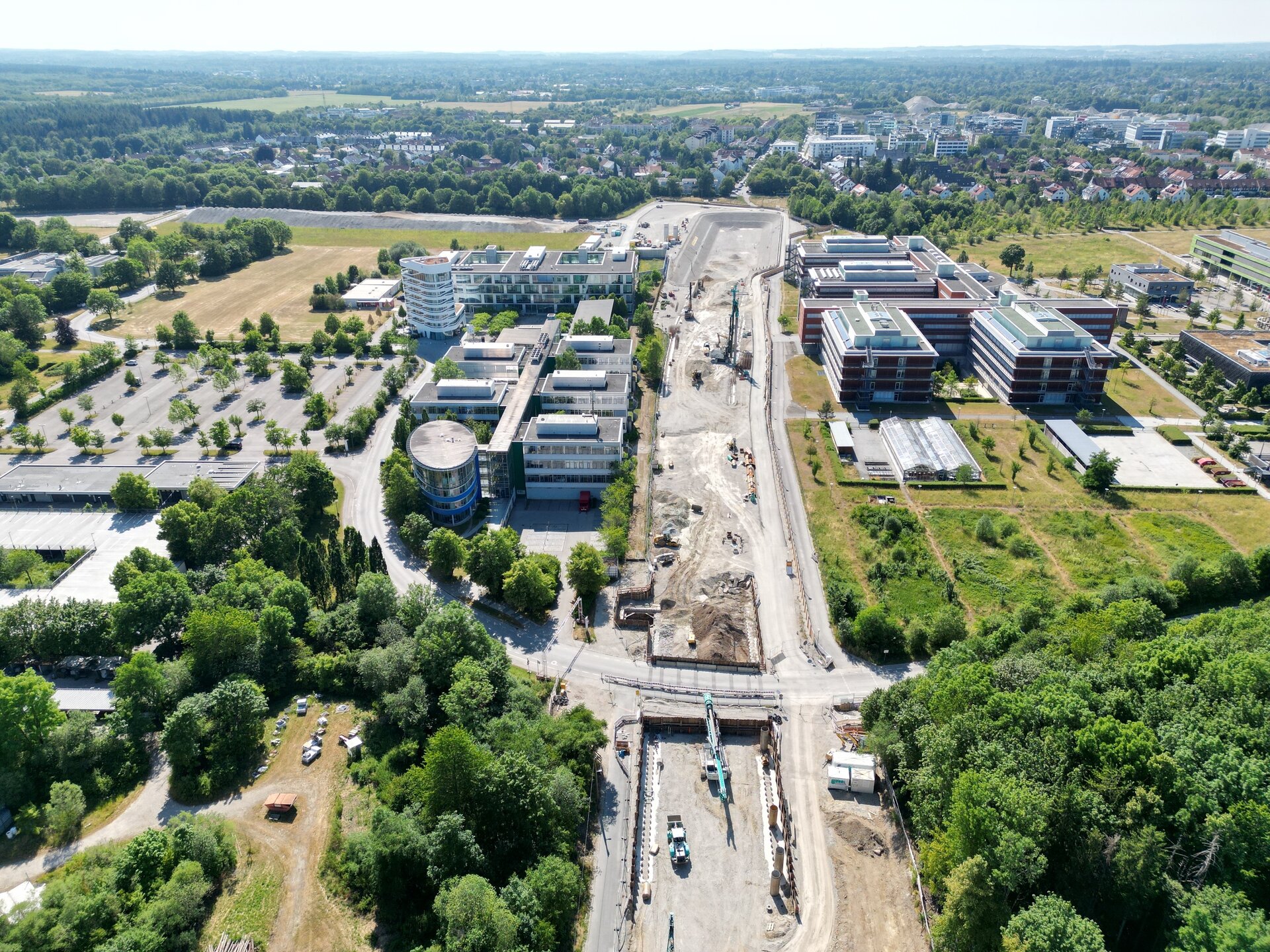Im Bild sind moderne Bürogebäude und eine Baustelle zu sehen, umgeben von viel Grün und einer Waldlandschaft im Hintergrund.