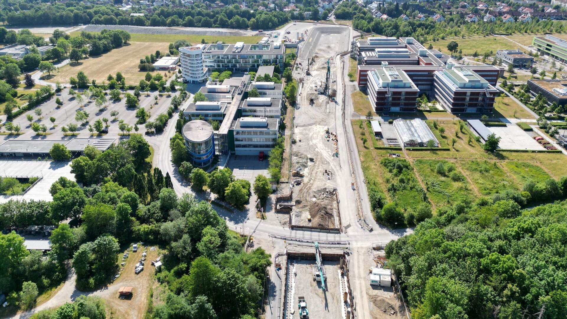 Im Bild sind moderne Bürogebäude und eine Baustelle zu sehen, umgeben von viel Grün und einer Waldlandschaft im Hintergrund.