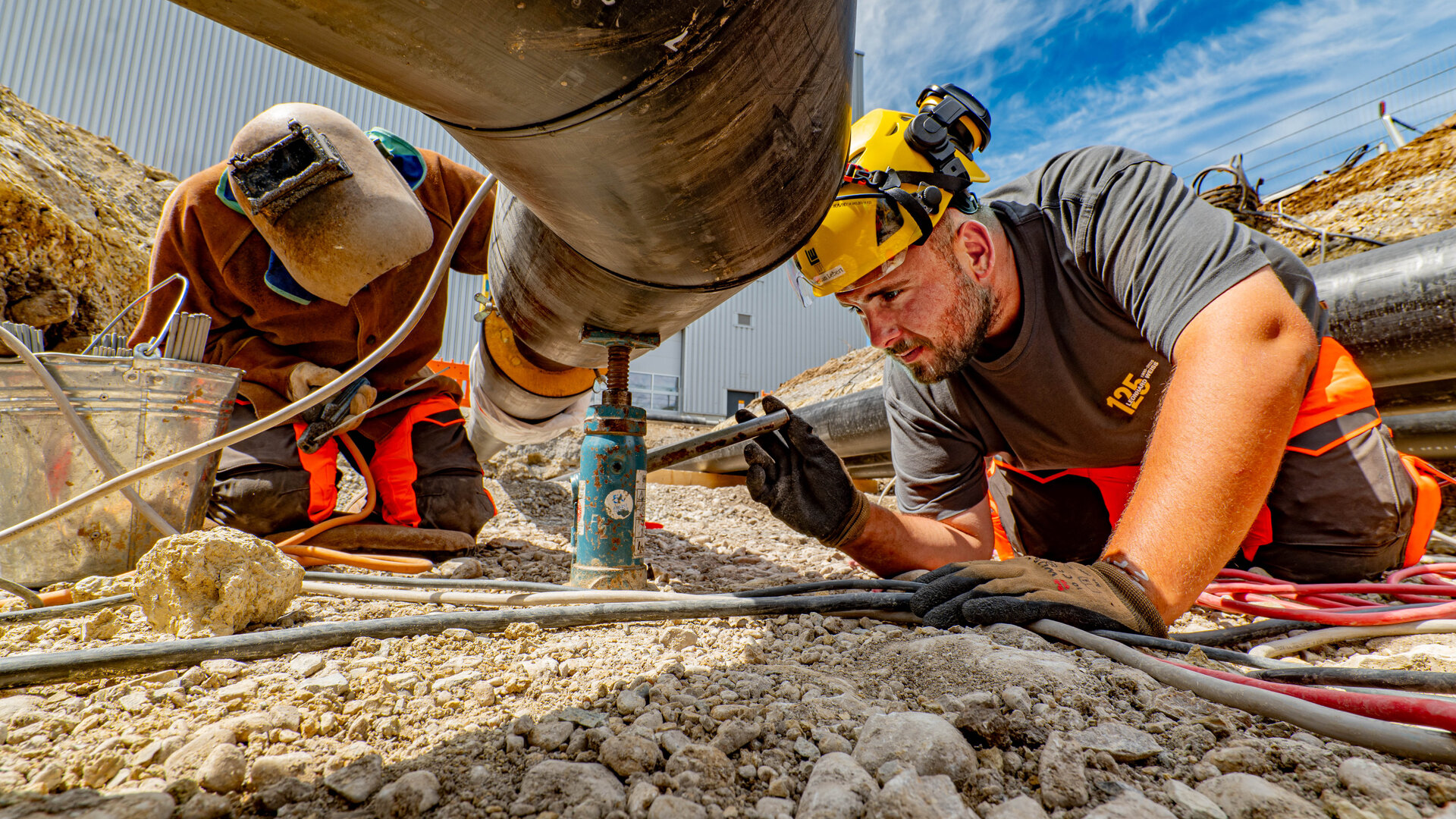 Ein Mann mit einem gelben Helm und Handschuhen führt Wartungsarbeiten an einer Rohrleitung durch, während ein Kollege im Hintergrund hilft.
