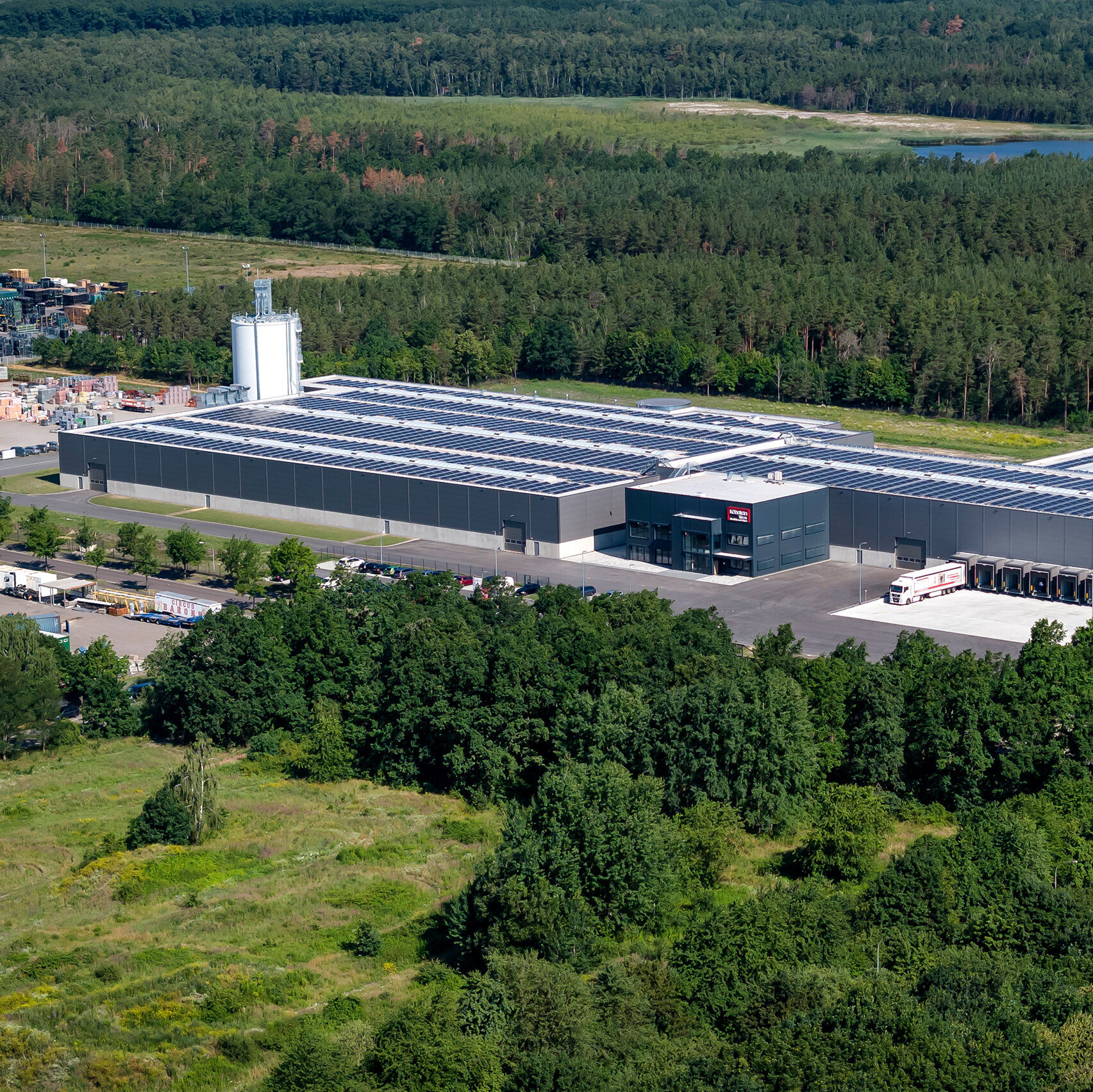 Eine moderne Lagerhalle mit Solarpanelen auf dem Dach, umgeben von dichten Wäldern und einem klaren See im Hintergrund.