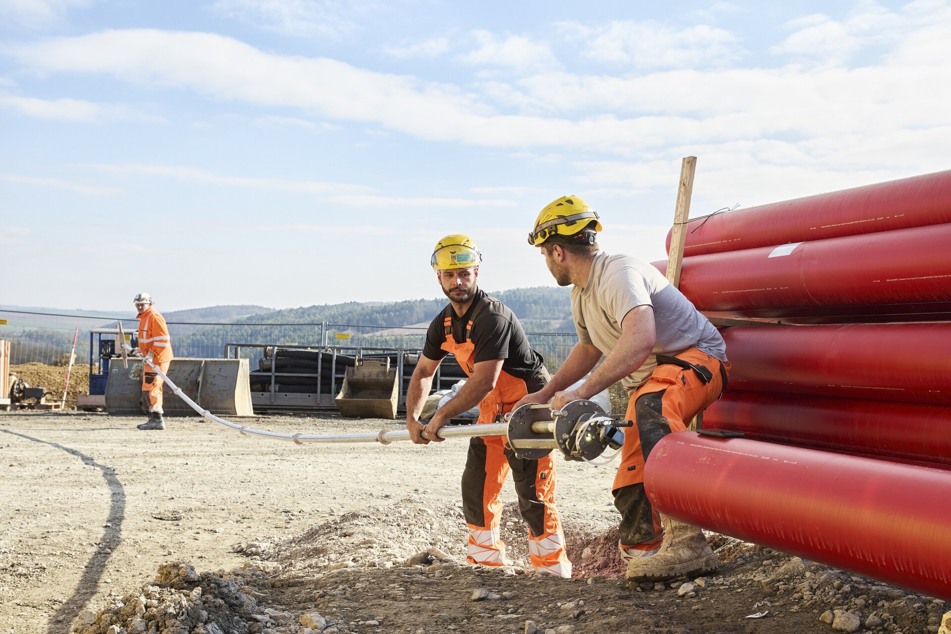 Die Szene zeigt zwei Bauarbeiter, die ein Rohr halten, während ein dritter Arbeiter im Hintergrund mit einem Werkzeug beschäftigt ist.