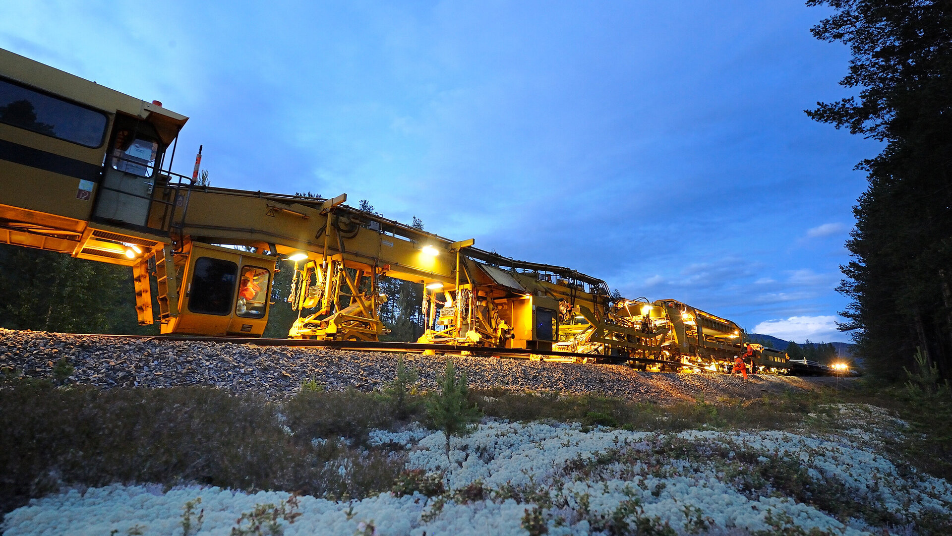 In der Dämmerung ist eine gelbe Gleisbaumaschine zu sehen, die mit Scheinwerfern die Schienen bearbeitet, umgeben von Natur.