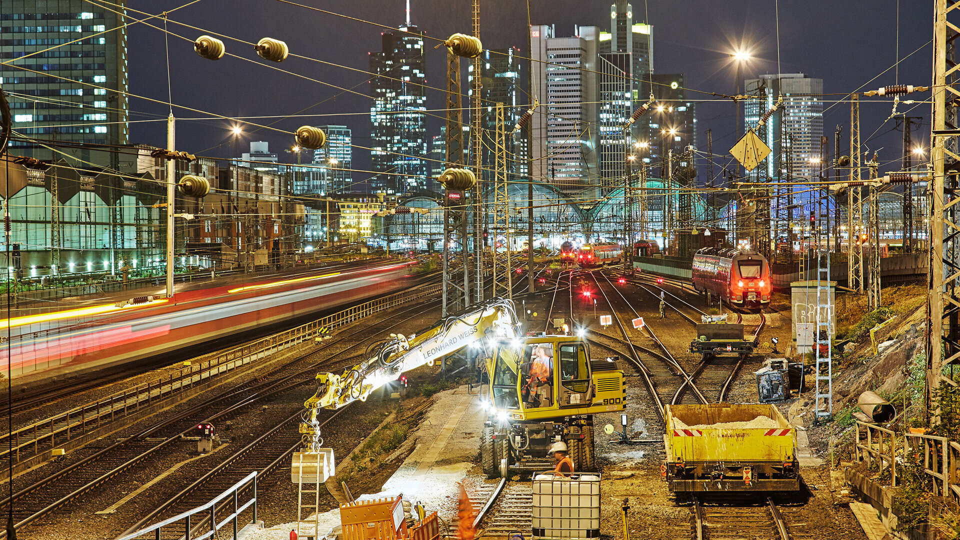 Im Hintergrund leuchten die Wolkenkratzer von Frankfurt, während ein Zug mit roten Lichtern an den Gleisen vorbeifährt.