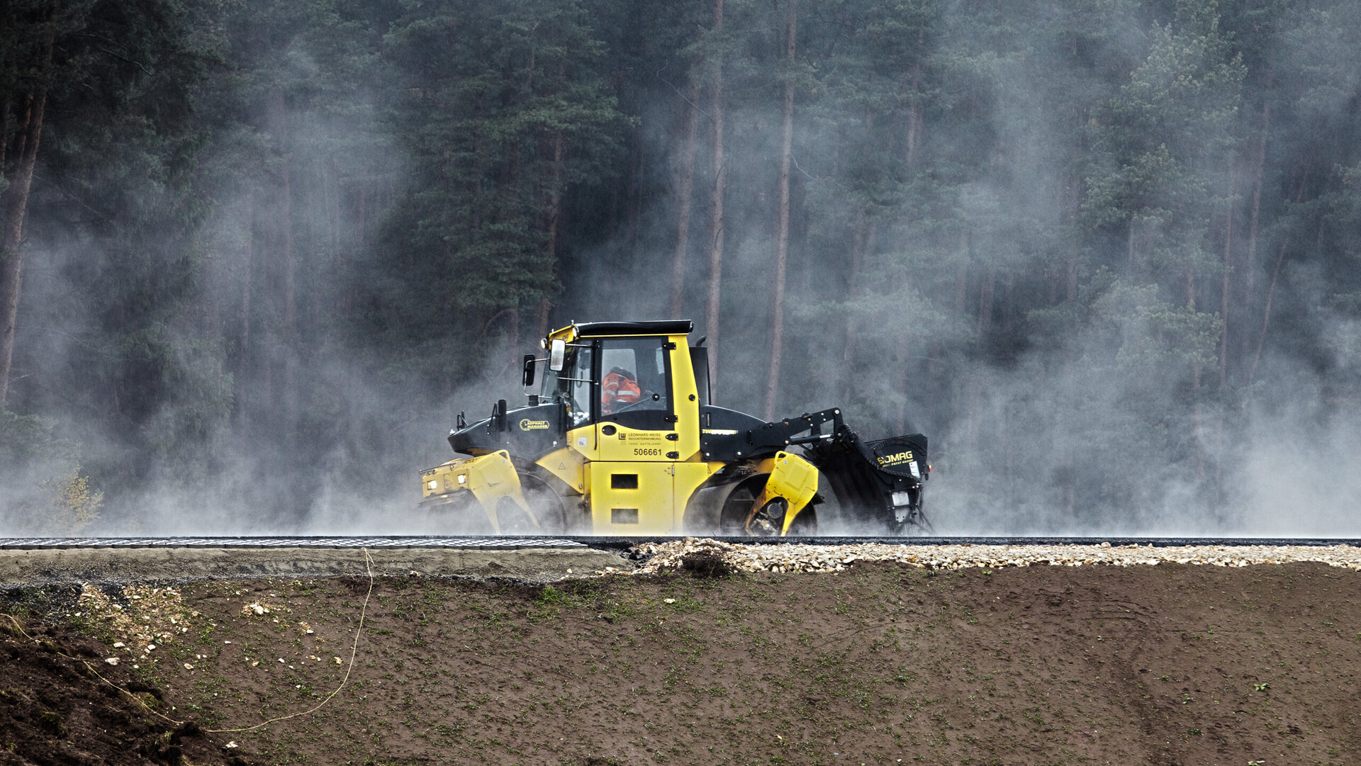 Die Maschine, umgeben von Dunst, bearbeitet den Boden, während Bäume im Hintergrund sichtbar sind und die Szene umrahmen.