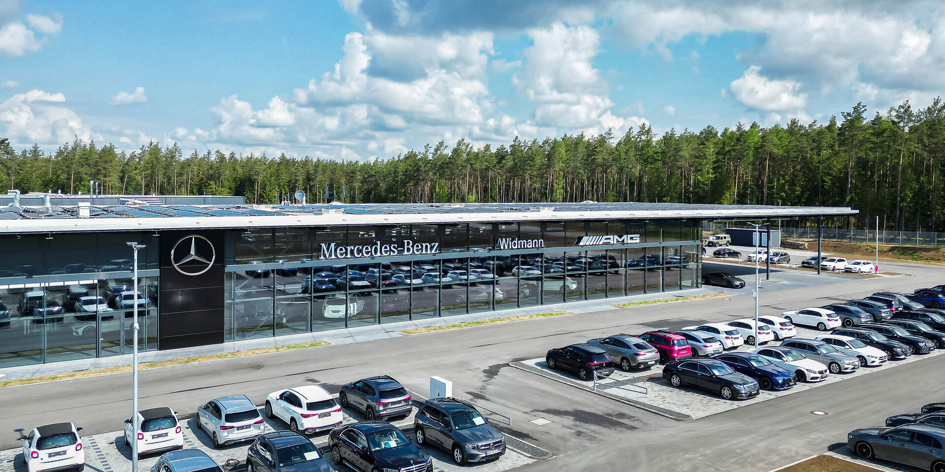Die Fassade eines Mercedes-Benz Autohauses zeigt zahlreiche Autos, während im Hintergrund grüne Bäume und Wolken sichtbar sind.