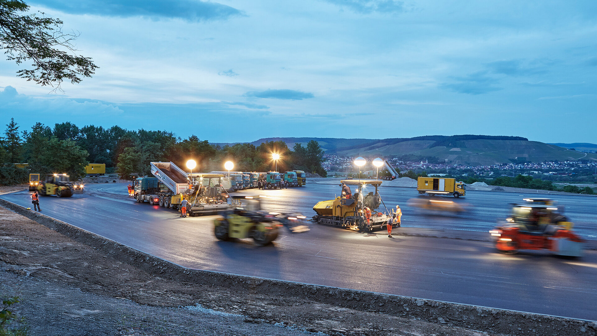 Bei Dämmerung arbeiten Bauarbeiter an einer neuen Straße, umgeben von schweren Maschinen und hellen Scheinwerfern.