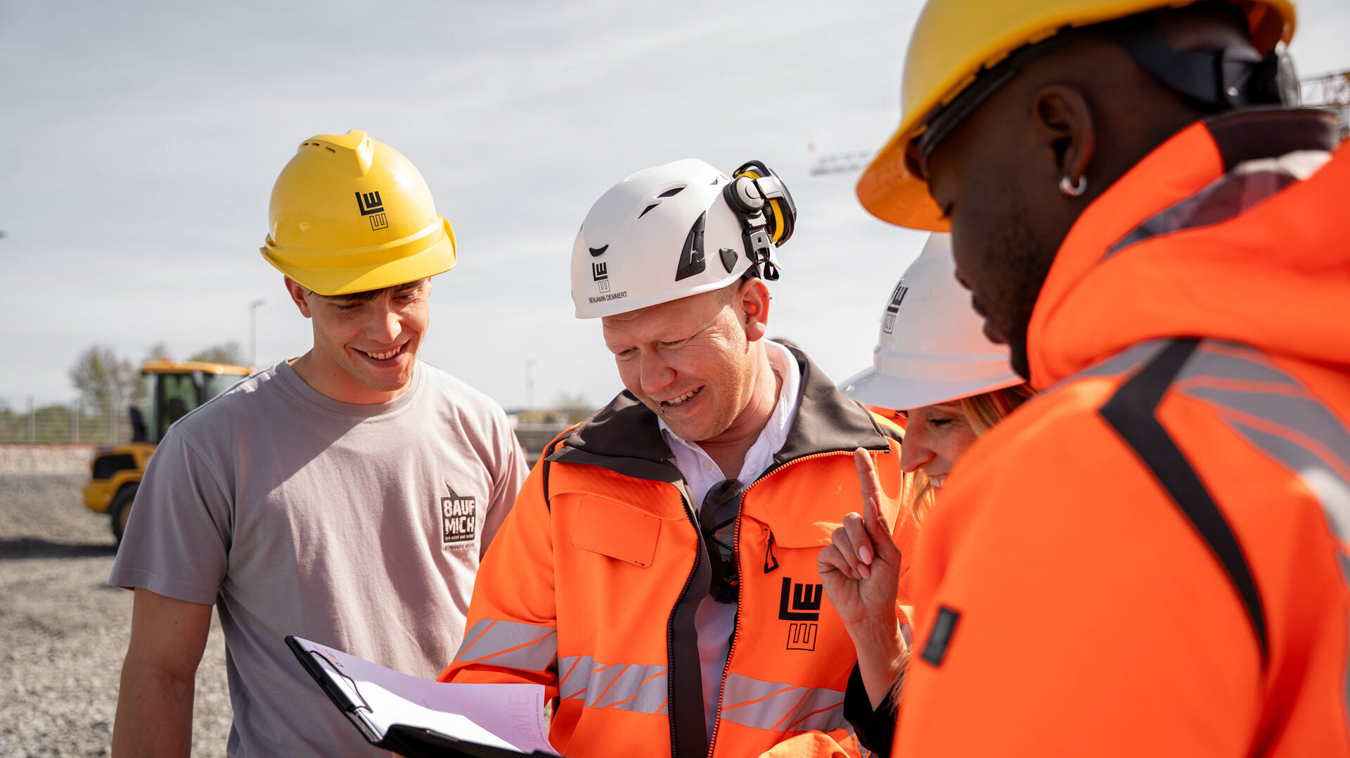 Ein Team von Bauprofis, gekleidet in leuchtend orangefarbene Jacken, analysiert gemeinsam ein Projekt auf einem sonnigen Baustellengelände.