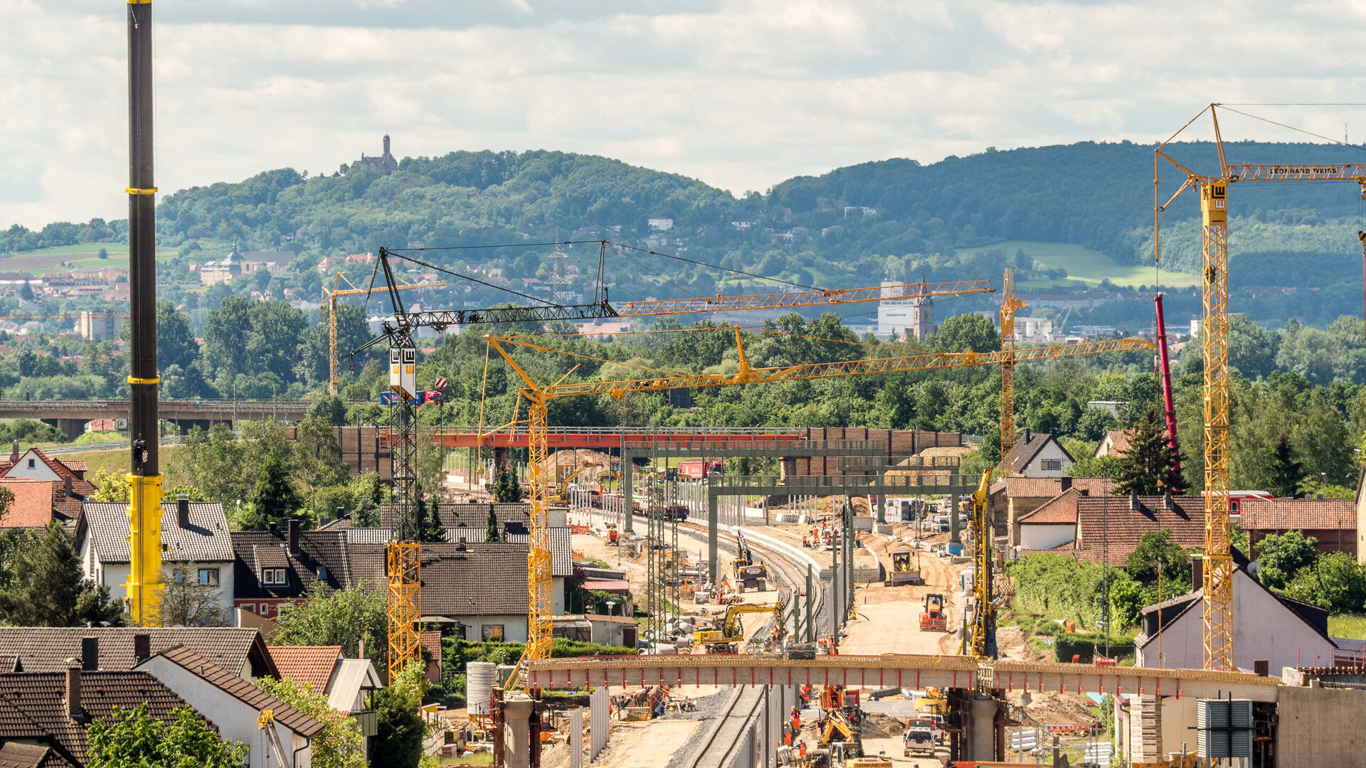 Eine Baustelle mit mehreren Kränen und Baufahrzeugen, die an einer neuen Eisenbahnlinie arbeiten, umgeben von grünen Hügeln.