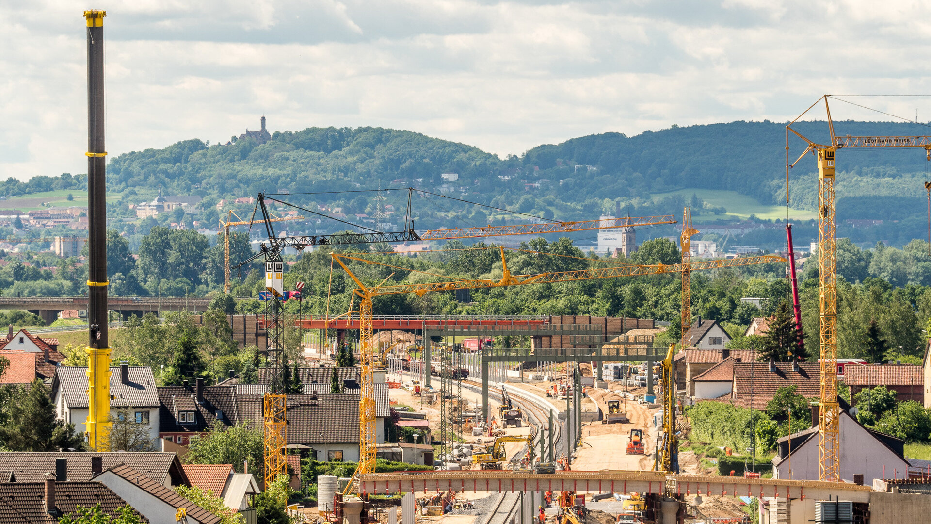 Eine Baustelle mit mehreren Kränen und Baufahrzeugen, die an einer neuen Eisenbahnlinie arbeiten, umgeben von grünen Hügeln.
