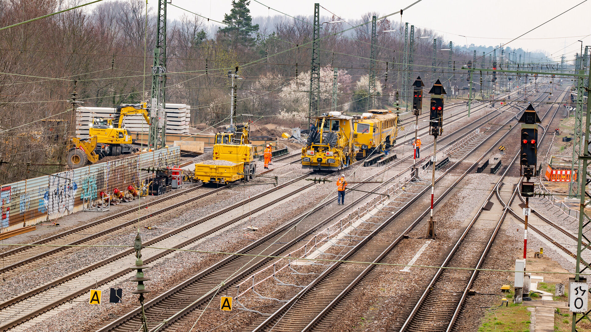 Auf einer Baustelle arbeiten mehrere gelbe Maschinen an den Gleisen, während Arbeiter in orangefarbener Kleidung aktiv sind.