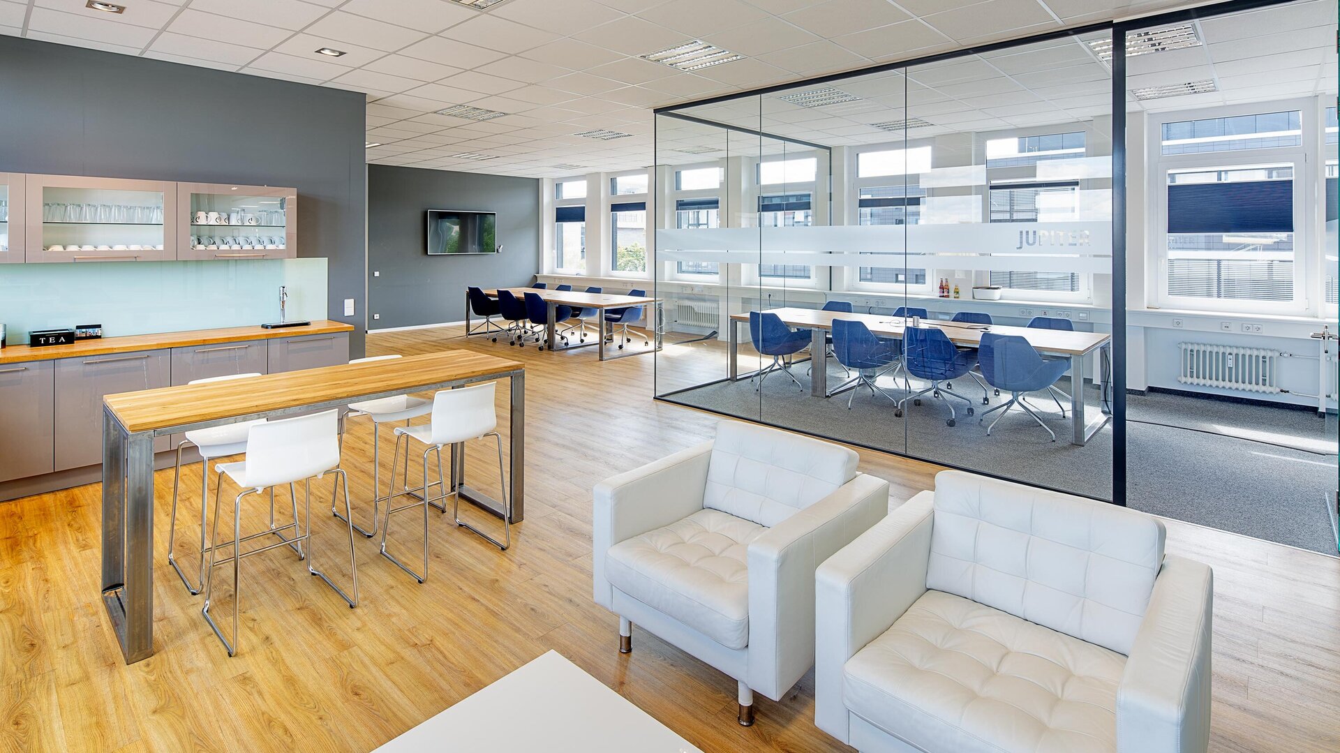 Ein modernes Büro mit einer hellen Küche, einem langen Tisch und blauen Stühlen, ideal für Besprechungen und Pausen.