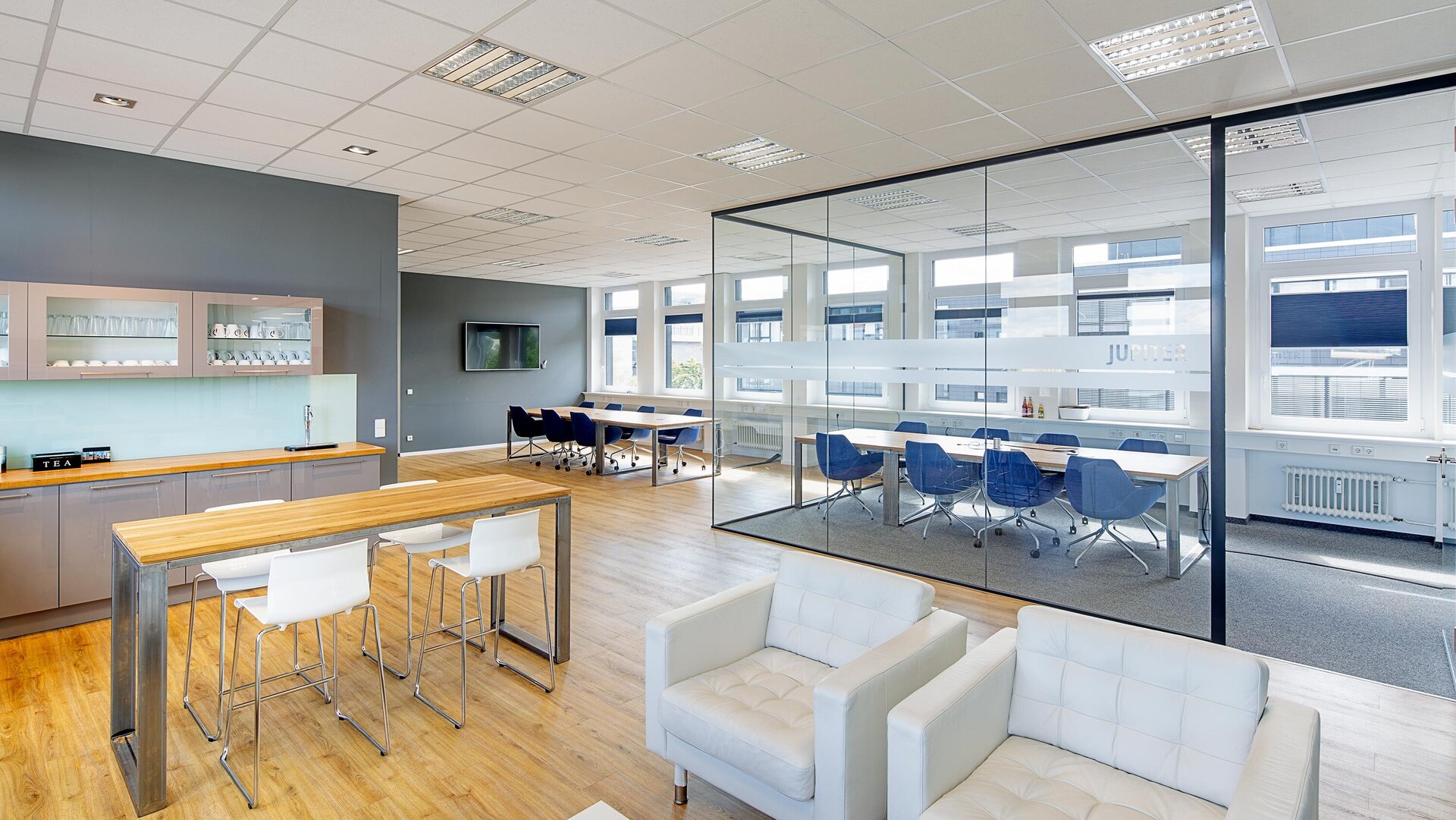 Ein modernes Büro mit einer hellen Küche, einem langen Tisch und blauen Stühlen, ideal für Besprechungen und Pausen.